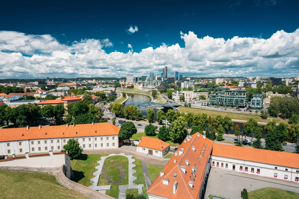 Wilno - idealne miasto na weekend Panorama miasta z widokiem na nowoczesne centrum z wysokimi budynkami, zielenią, rzeką płynącą przez środek oraz historycznymi budynkami z czerwonymi dachami na pierwszym planie.