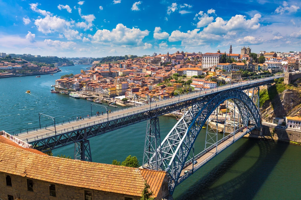 Tanie wakacje w Porto Most z łukowym przęsłem łączy dwa brzegi rzeki wśród gęsto zabudowanej miejskiej panoramy, po jednej stronie widoczne dachy czerwonych budynków oraz zabytkowe kościoły, w tle rozciąga się malownicza sceneria z błękitnym niebem i chmurami.