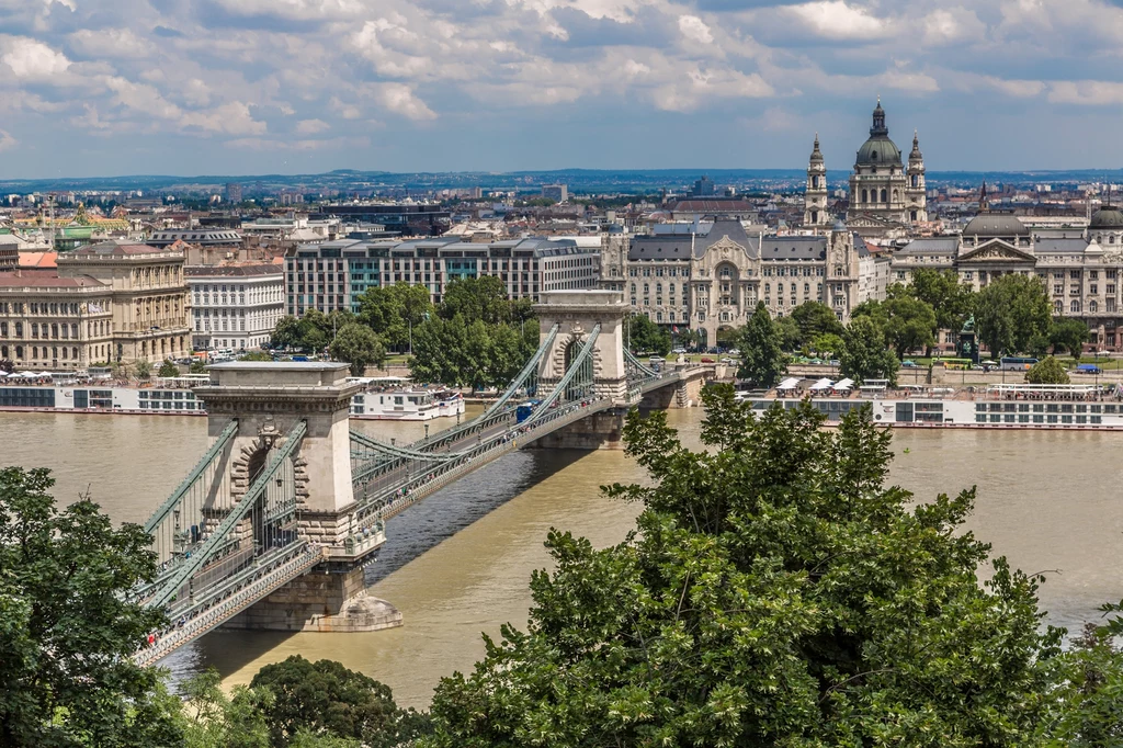 Budapeszt Most Łańcuchowy na Dunaju łączący dwa brzegi Budapesztu, otoczony historycznymi budynkami oraz zielenią w pierwszym planie, w tle panorama miasta z charakterystyczną bazyliką.