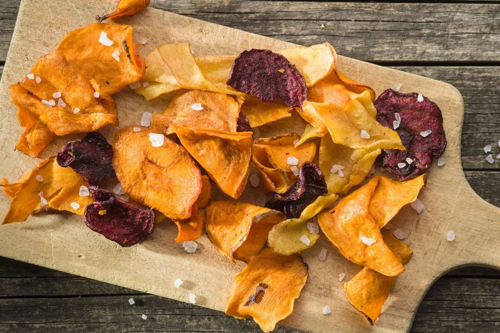 Przygotuj z rzepy, buraka, marchwi i pietruszki pyszne chipsy z piekarnika Kolorowe chipsy warzywne z batata, buraka i marchewki posypane gruboziarnistą solą leżą na drewnianej desce, kontrastując z fakturą starego drewnianego stołu.