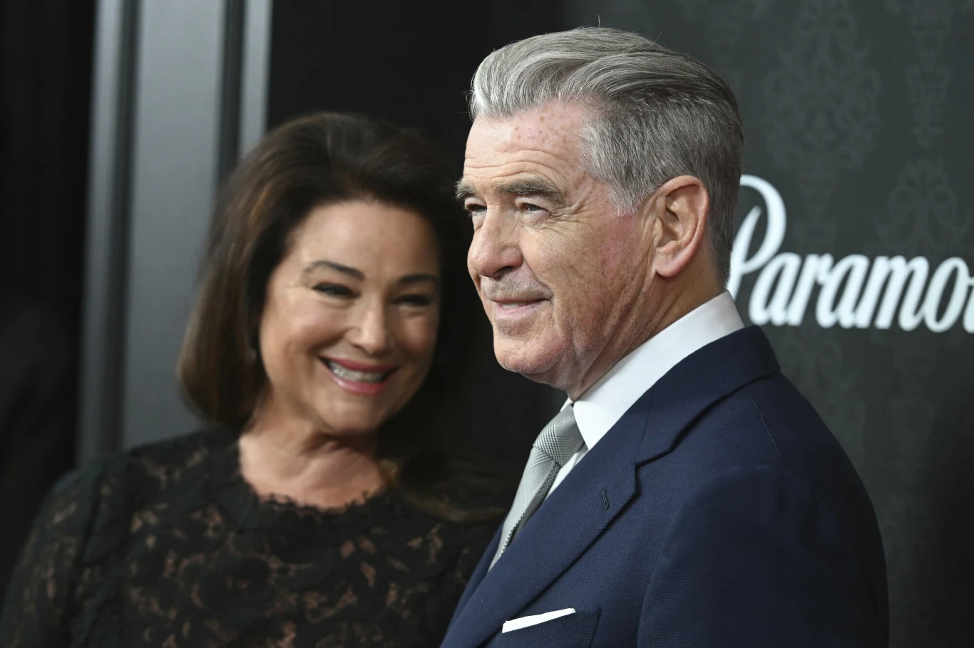 Keely Shaye Smith i Pierce Brosnan przyciągnęli wzrok zebranych Uśmiechnięta kobieta stojąca obok mężczyzny w eleganckim garniturze na tle dekoracyjnej ściany z logo Paramount.
