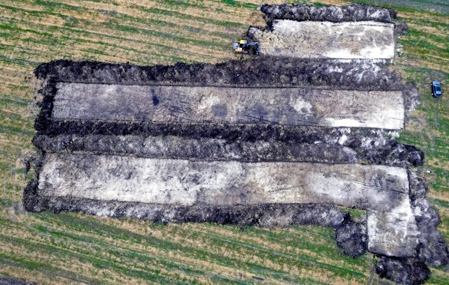 Odhumusowany teren badań, widok z drona / Lubelski Wojewódzki Konserwator Zabytków (fot. P. Szelest). Wykopaliska archeologiczne ukazujące trzy równoległe, długie wykopy na zielonym polu z widocznymi fragmentami ziemi oraz sprzętem wykorzystywanym do prac terenowych.