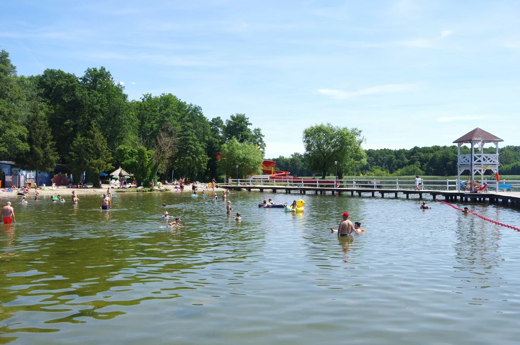 Wakacje na Pojezierzu Lubuskim? Jak najbardziej! Plaża nad jeziorem z wieloma kąpiącymi się osobami, pomostem z barierkami i białą altaną, otoczona drzewami pod błękitnym niebem.