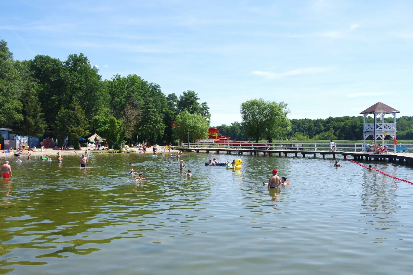 Plaża nad jeziorem z wieloma kąpiącymi się osobami, pomostem z barierkami i białą altaną, otoczona drzewami pod błękitnym niebem.