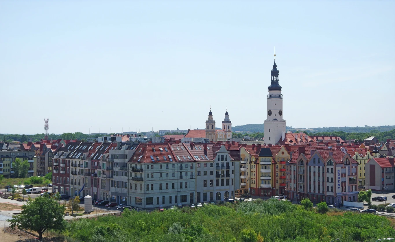 Zabudowa miejska z rzędem kolorowych kamienic o czerwonych dachach, w tle góruje wysoki kościół z charakterystyczną wieżą, otoczenie zieleni i niskiej zabudowy mieszkalnej