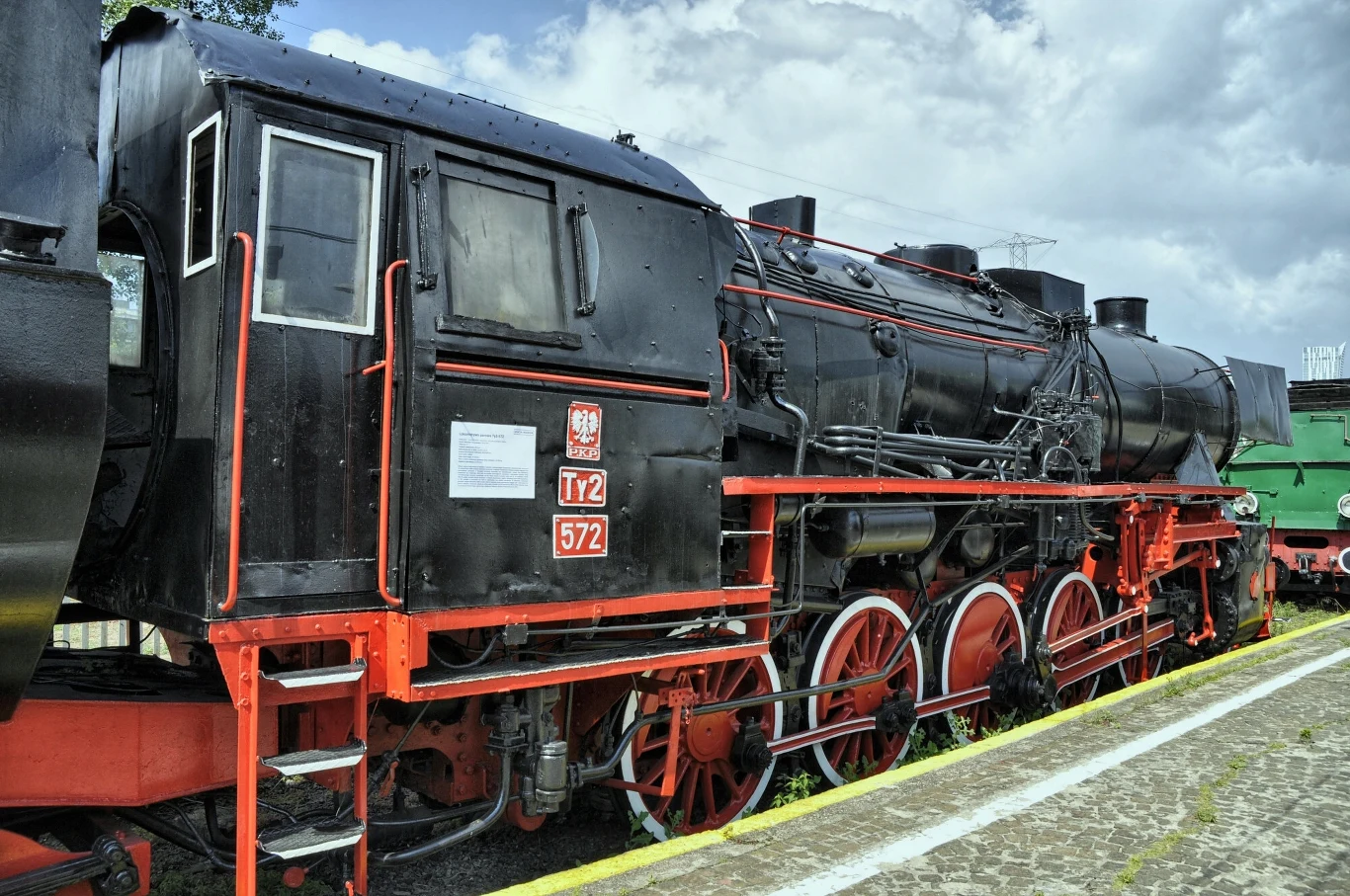 Podróż w historycznych wagonach, ciągniętych przez zabytkową lokomotywę to wyjątkowa atrakcja Czarna lokomotywa parowa typu Ty2 z czerwonymi elementami i dużymi kołami stoi na torze kolejowym obok brukowanego peronu, w tle widoczny fragment kolejnej lokomotywy oraz niebo z chmurami.