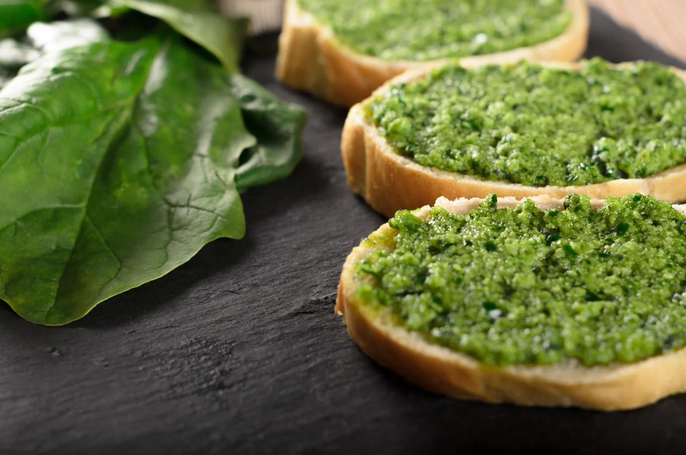 Z mniszka lekarskiego warto przygotować smaczne i zdrowe pesto Grzanki z chrupiącego chleba posmarowane zielonym pesto, ułożone na ciemnym kamieniu, obok świeże liście szpinaku.