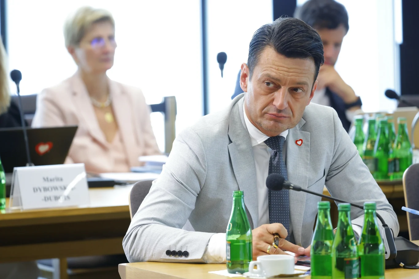 Kontrowersyjne słowa Przemysława Witka. Czy pogrążą Trzaskowskiego? Mężczyzna w jasnym garniturze siedzi przy stole konferencyjnym, przed nim stoi mikrofon i kilka butelek wody, w tle inni uczestnicy spotkania, atmosfera formalna.