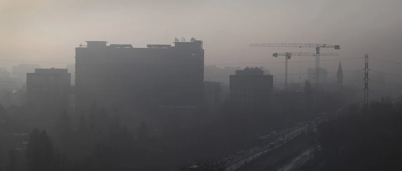 Budynki i żurawie budowlane ledwo widoczne przez gęsty smog pokrywający miasto oraz znaczną część widnokręgu, w oddali ledwo widoczne sylwetki dachów i wieży.