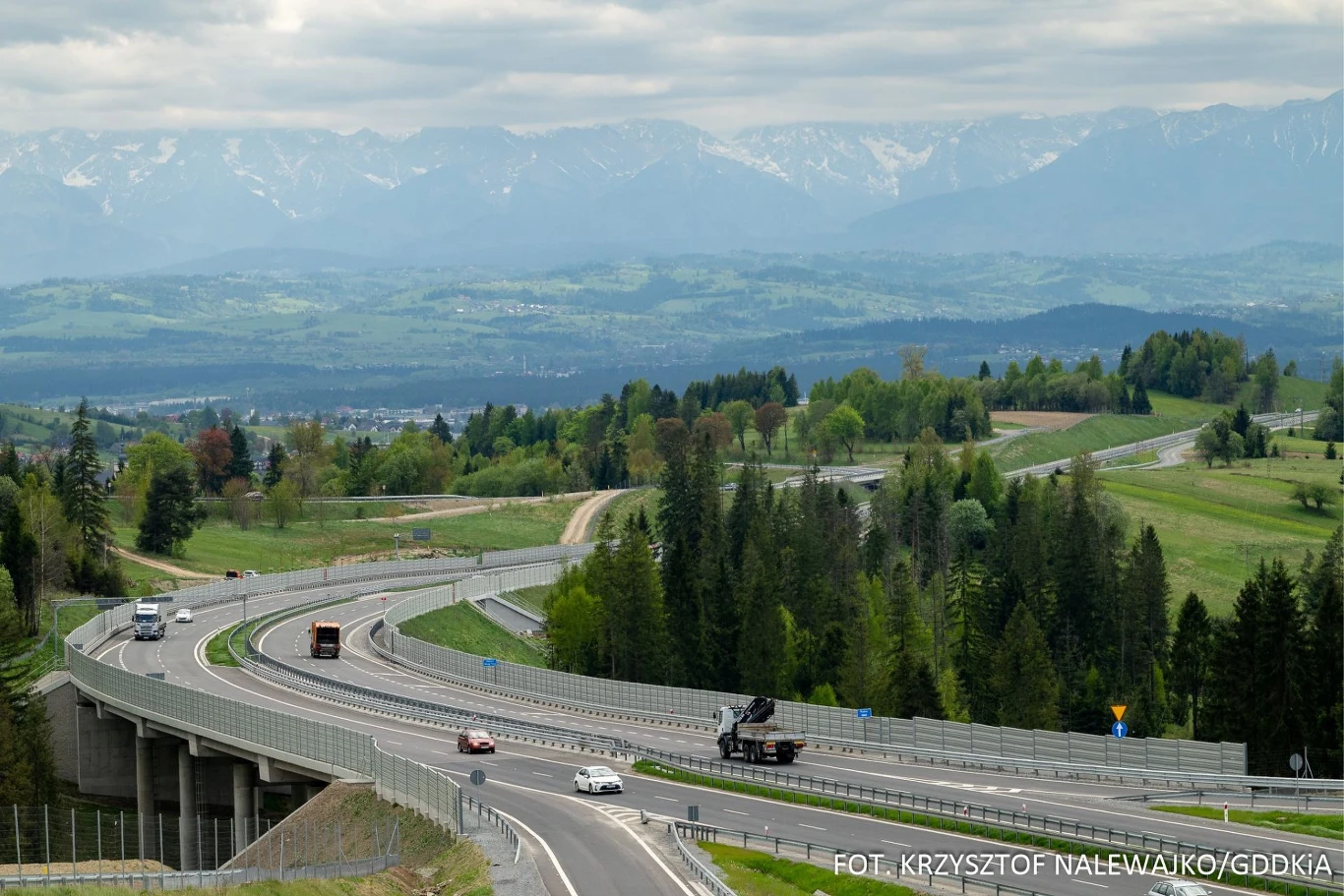 Nowoczesna DK47 w wiosennej scenerii prezentuje się wyjątkowo urokliwie. nowoczesna droga ekspresowa wijąca się przez zielone wzgórza, otoczona drzewami i lasami, z pojazdami jadącymi w obu kierunkach, w tle rozciąga się malowniczy widok na górskie pasma pokryte mgłą