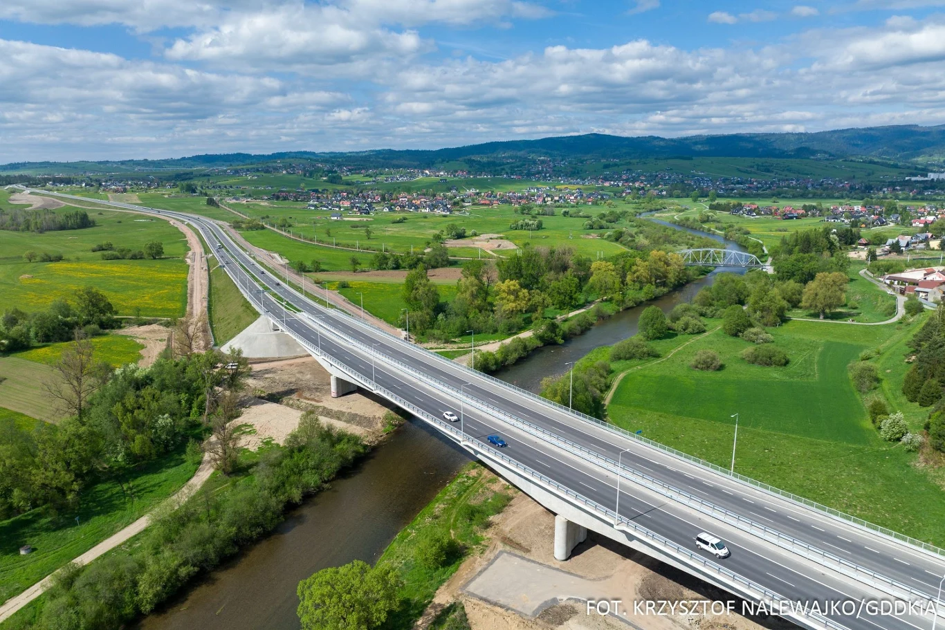 DK47 to drogowy majstersztyk idealnie wkomponowany w naturę. Most drogowy przebiegający nad rzeką na tle rozległej, zielonej doliny oraz otoczenia zabudowań wiejskich; widoczne samochody przemieszczające się po nowoczesnej konstrukcji, krajobraz z licznymi polami i wzgórzami w tle.