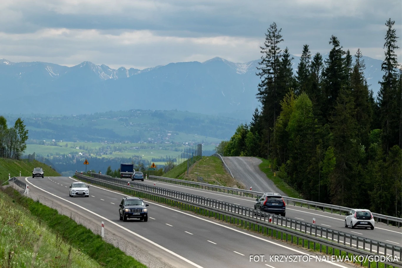 Przez wielu jest określana najpiękniejszą drogą w Polsce. Nowoczesna droga ekspresowa prowadząca przez malowniczy, górski teren, wokół zielone lasy i rozległa dolina, w tle widoczne ośnieżone szczyty gór oraz chmury na niebie, po obu pasach drogi poruszają się samochody.