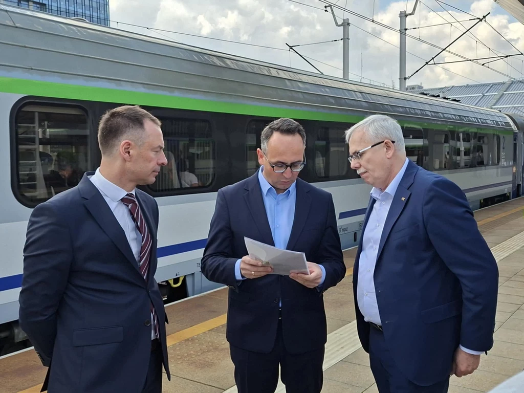 O szczegółach pociągu do Chorwacji mówili (od prawej) prezes PKP Intercity Janusz Malinowski, minister infrastruktury Dariusz Klimczak oraz wiceszef resortu sportu i turystyki Piotr Borys Trzech elegancko ubranych mężczyzn w garniturach stoi na peronie obok nowoczesnego pociągu; jeden z nich trzyma dokumenty i uważnie je przegląda, pozostali przyglądają się z zainteresowaniem.