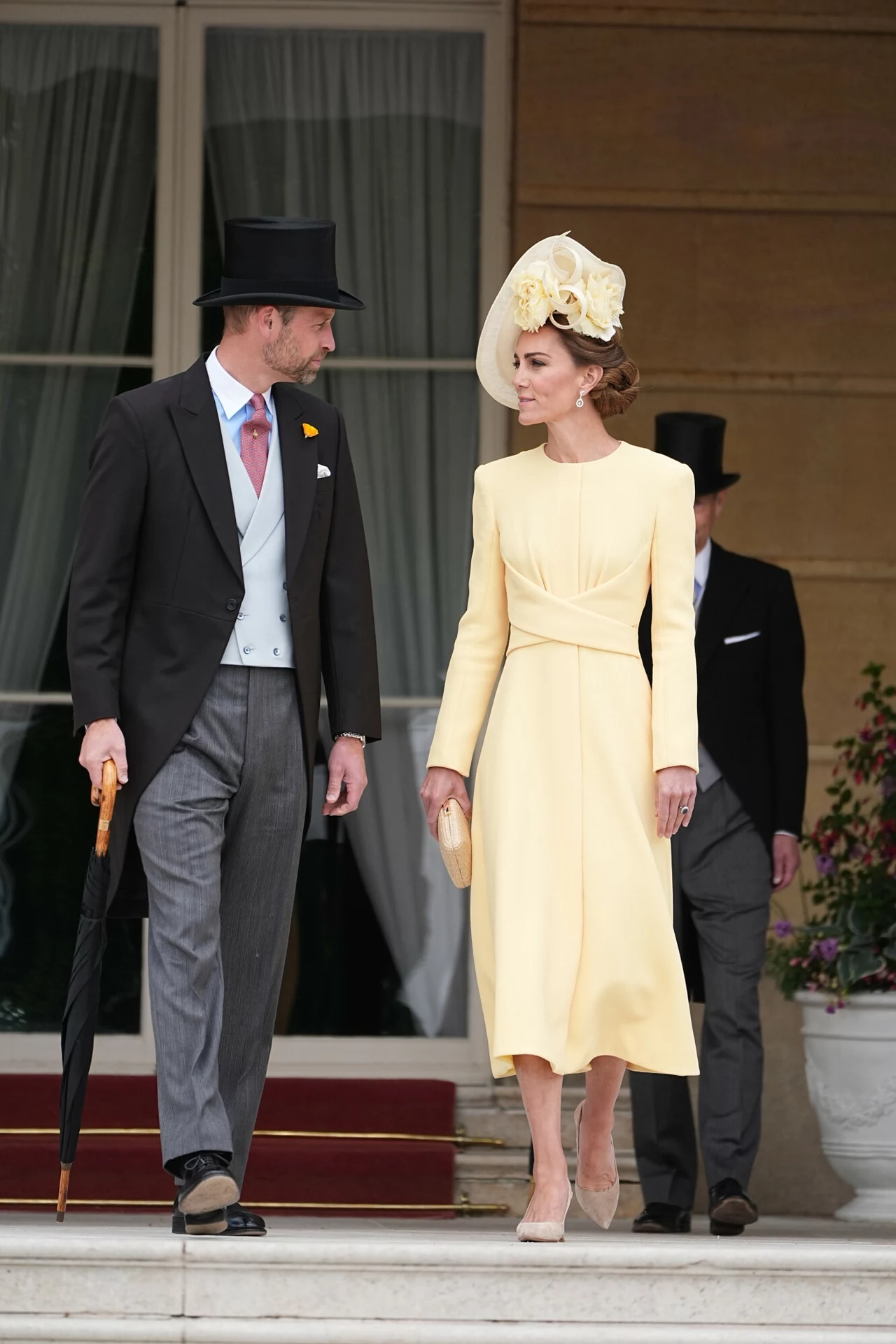 Księżna Kate i książę William Elegancko ubrany mężczyzna w cylindrze rozmawia z kobietą w jasnożółtej sukience i fantazyjnym kapeluszu, oboje stoją na marmurowych schodach przed wejściem do budynku z dużymi oknami, w tle widoczna jest jeszcze jedna osoba w podobnym stroju formalnym.
