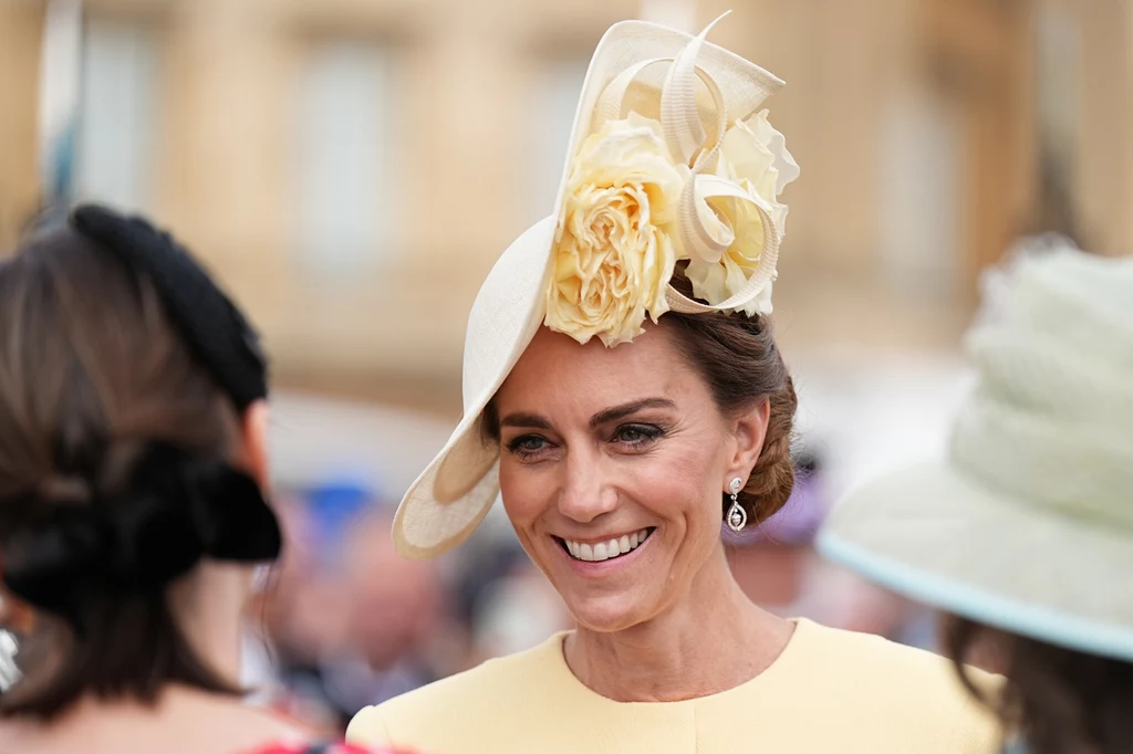 Księżna Kate w oryginalnym fascynatorze Elegancko ubrana kobieta w kremowym kapeluszu ozdobionym dużymi kwiatami w odcieniach żółci, uśmiechnięta, rozmawia z innymi osobami w tle, które są lekko rozmazane.