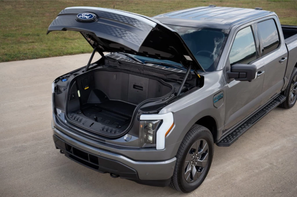 Zamiast potężnego silnika V8 pod maską F-150 Lightning znajduje się dodatkowa przestrzeń bagażowa. Srebrny pick-up Ford z otwartą przednią maską, ukazującą pojemny bagażnik w miejscu tradycyjnego silnika, pojazd stoi na utwardzonej nawierzchni, wokół trawiasta okolica.