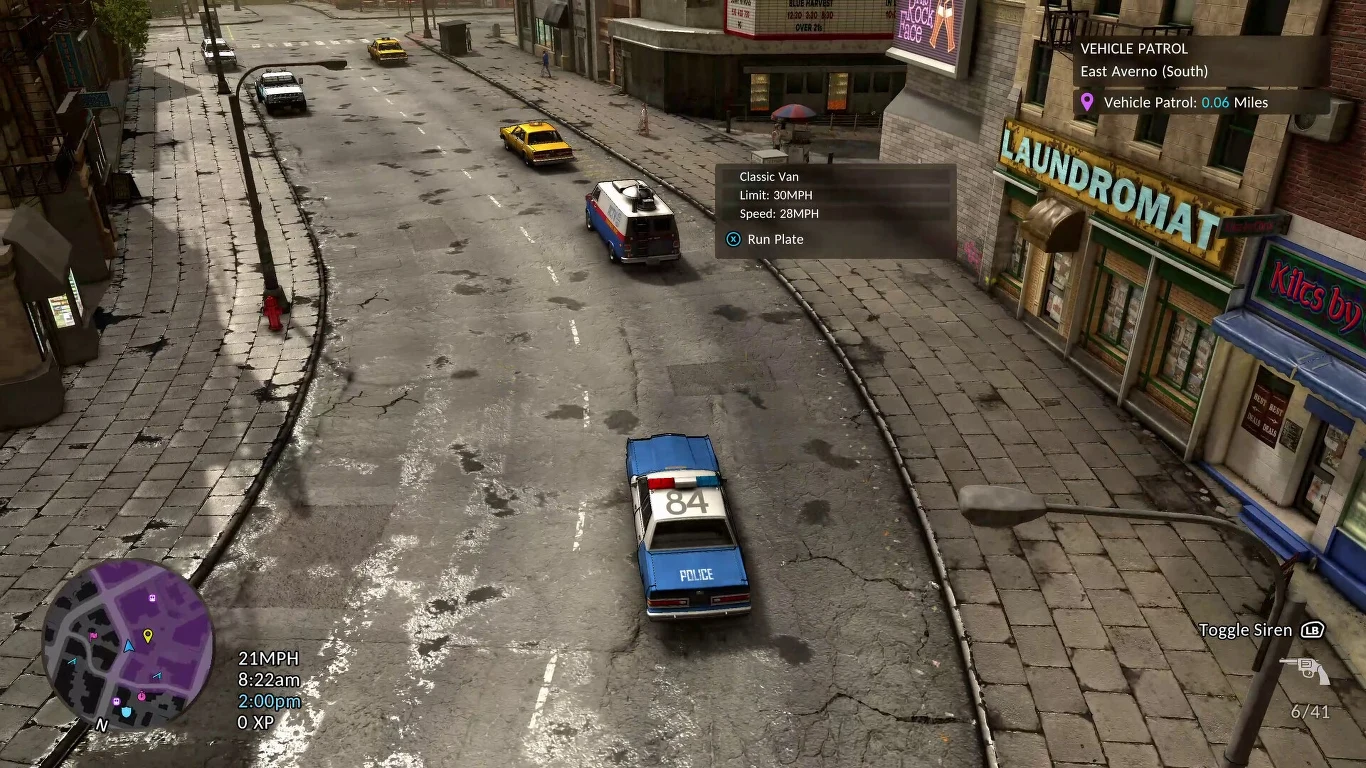 The Precinct - screen z gry Pusta, lekko zaniedbana ulica miejska, po której poruszają się samochody w tym radiowóz policyjny widoczny na środku, budynki z szyldami sklepów, w tym pralnia po prawej stronie.