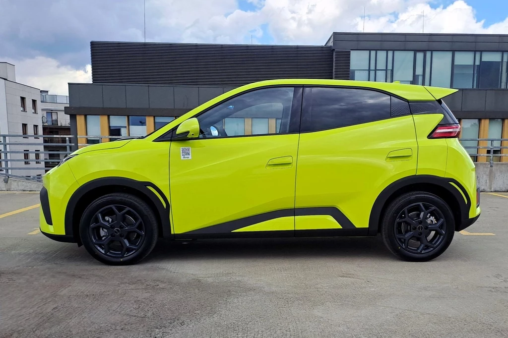 Niespełna czterometrowy BYD Dolphin Surf ma rozstaw osi na poziomie 2,5 metra. jasnozielony samochód elektryczny o nowoczesnych, dynamicznych liniach nadwozia, stojący na betonowym parkingu, w tle miejska zabudowa i nowoczesne biurowce