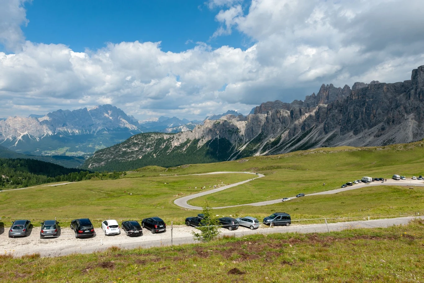 Kręta asfaltowa droga na tle rozległych, zielonych łąk prowadzi wśród górskich szczytów i skalistych zboczy, na poboczu stoi rząd zaparkowanych samochodów, nad całością unoszą się białe chmury na niebieskim niebie.