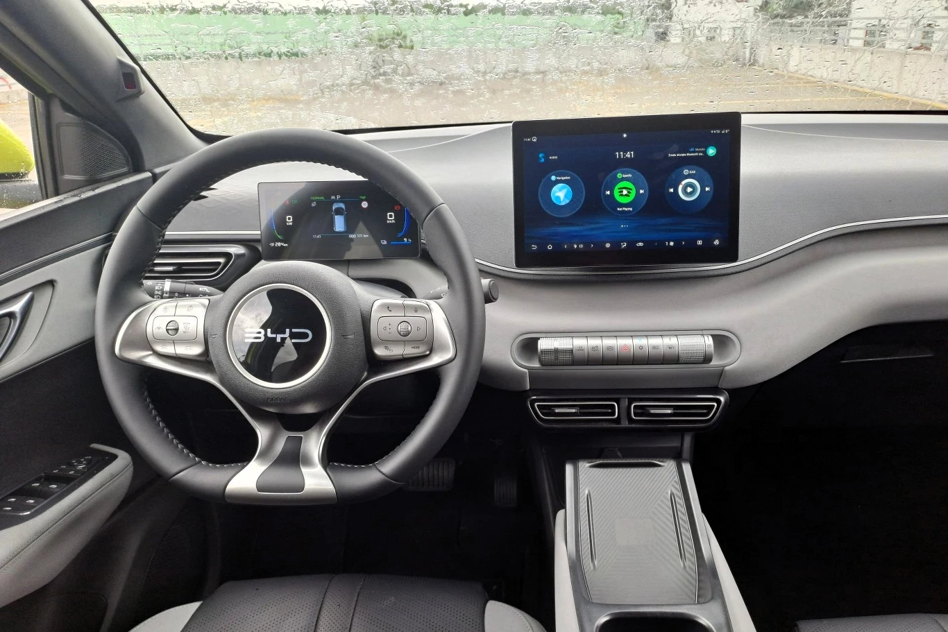 Nowoczesna deska rozdzielcza samochodu elektrycznego z kierownicą z logo BYD, dużym, dotykowym ekranem systemu multimedialnego po prawej stronie, cyfrowym zegarami przed kierowcą, minimalistycznymi przyciskami oraz wygodnym podłokietnikiem.