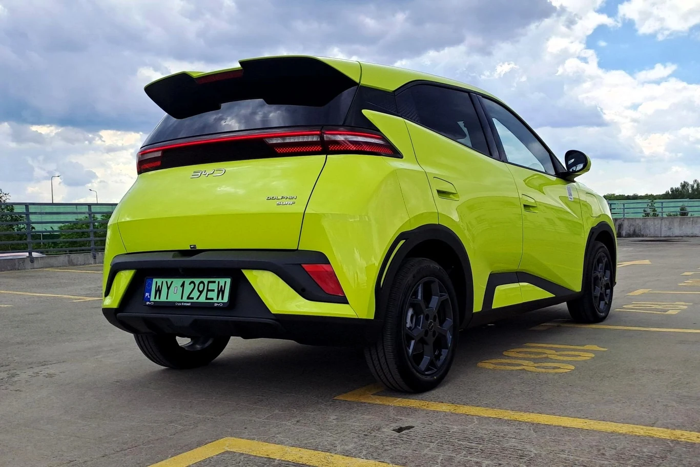 Jaskrawozielony samochód osobowy marki BYD Dolphin stoi na parkingu wielopoziomowym, na tle zachmurzonego nieba, widoczny tył auta z charakterystycznymi czarnymi akcentami i sportowym spojlerem.