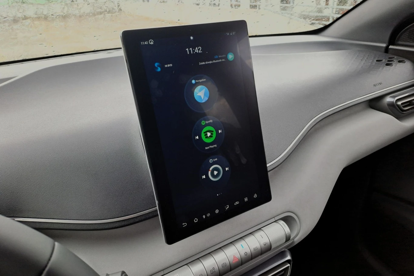 Duży dotykowy ekran multimedialny zamontowany na konsoli środkowej w nowoczesnym samochodzie, z wyświetlonymi ikonami nawigacji, muzyki i telefonu, wokół widoczne elementy minimalistycznego kokpitu.