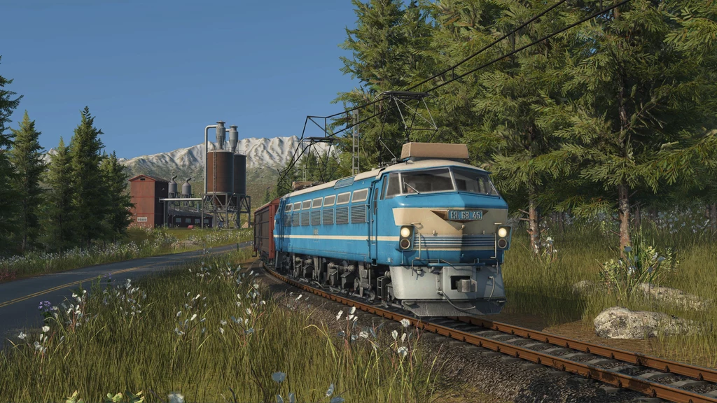 Transport Fever 3 - screen z gry Lokomotywa elektryczna poruszająca się po torach, otoczona bujną zielenią i wysokimi drzewami iglastymi, w tle widoczna fabryka i ośnieżone szczyty gór.