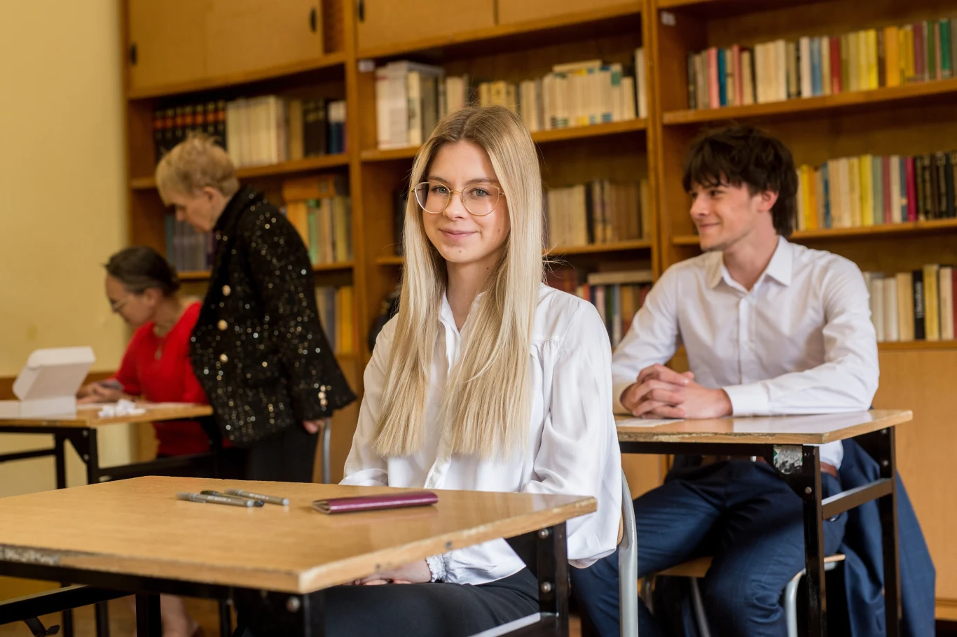 Matura 2025 już za uczniami Uczniowie siedzący w ławkach w klasie, na pierwszym planie uśmiechnięta młoda dziewczyna w okularach, w tle pozostali uczniowie i nauczycielki w sali z regałem pełnym książek.