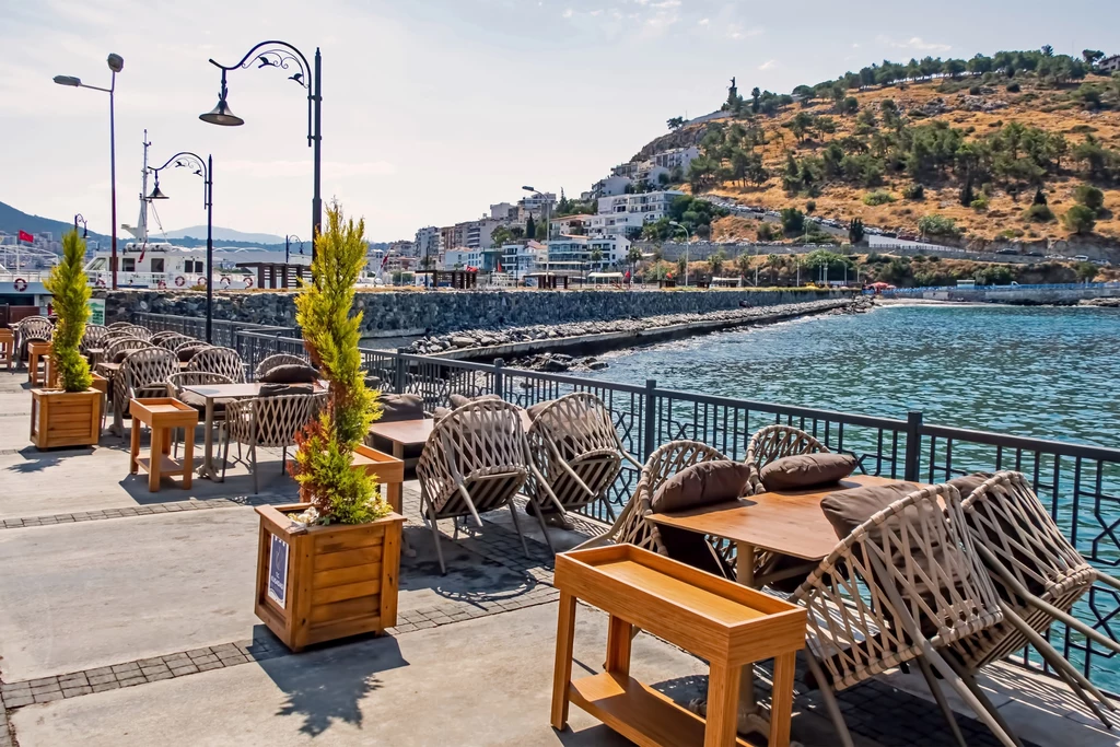 Jednym z miasteczek, które warto odwiedzić w regionie Morza Egejskiego jest Kuşadası Pusty taras restauracyjny nad brzegiem morza z wiklinowymi krzesłami i drewnianymi stołami, ozdobiony donicami z roślinami, w tle nadmorska promenada, wzgórze porośnięte krzewami oraz budynki mieszkalne.