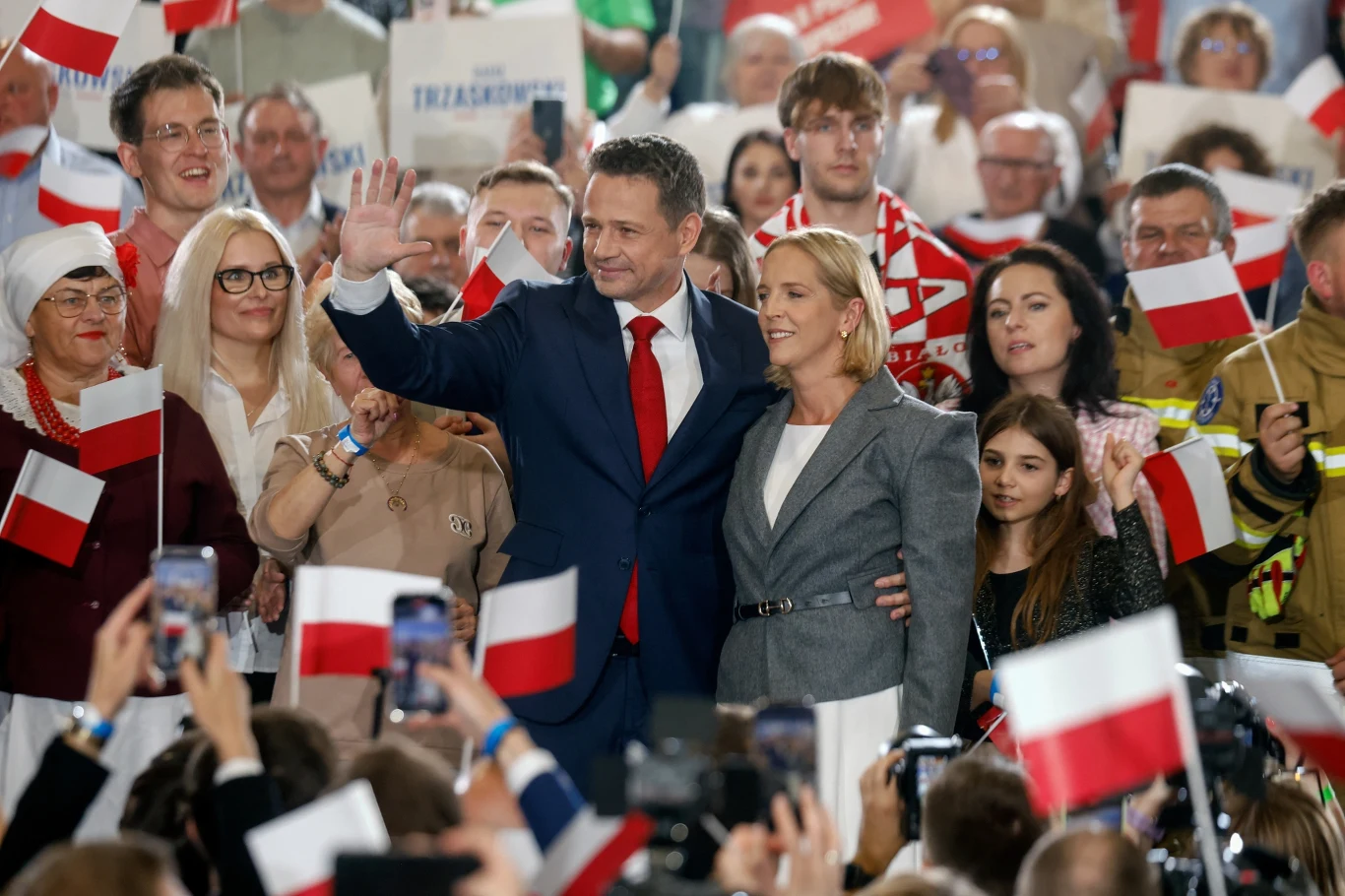 Polityk w garniturze z czerwoną krawatą wita się z tłumem, obok stoi kobieta i młoda dziewczyna, wokół zgromadzeni ludzie z flagami Polski, w tle widoczne transparenty oraz osoby w różnym wieku.