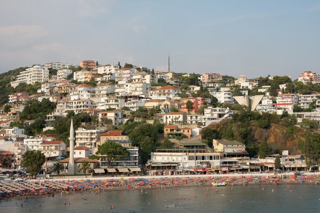 Ulcinj - miasto na południu Czarnogóry Plaża pełna ludzi nad morzem, na pierwszym planie widać parasole i kąpiących się, w tle wzgórze gęsto zabudowane białymi budynkami mieszkalnymi, z widocznym minaretem meczetu po lewej stronie.