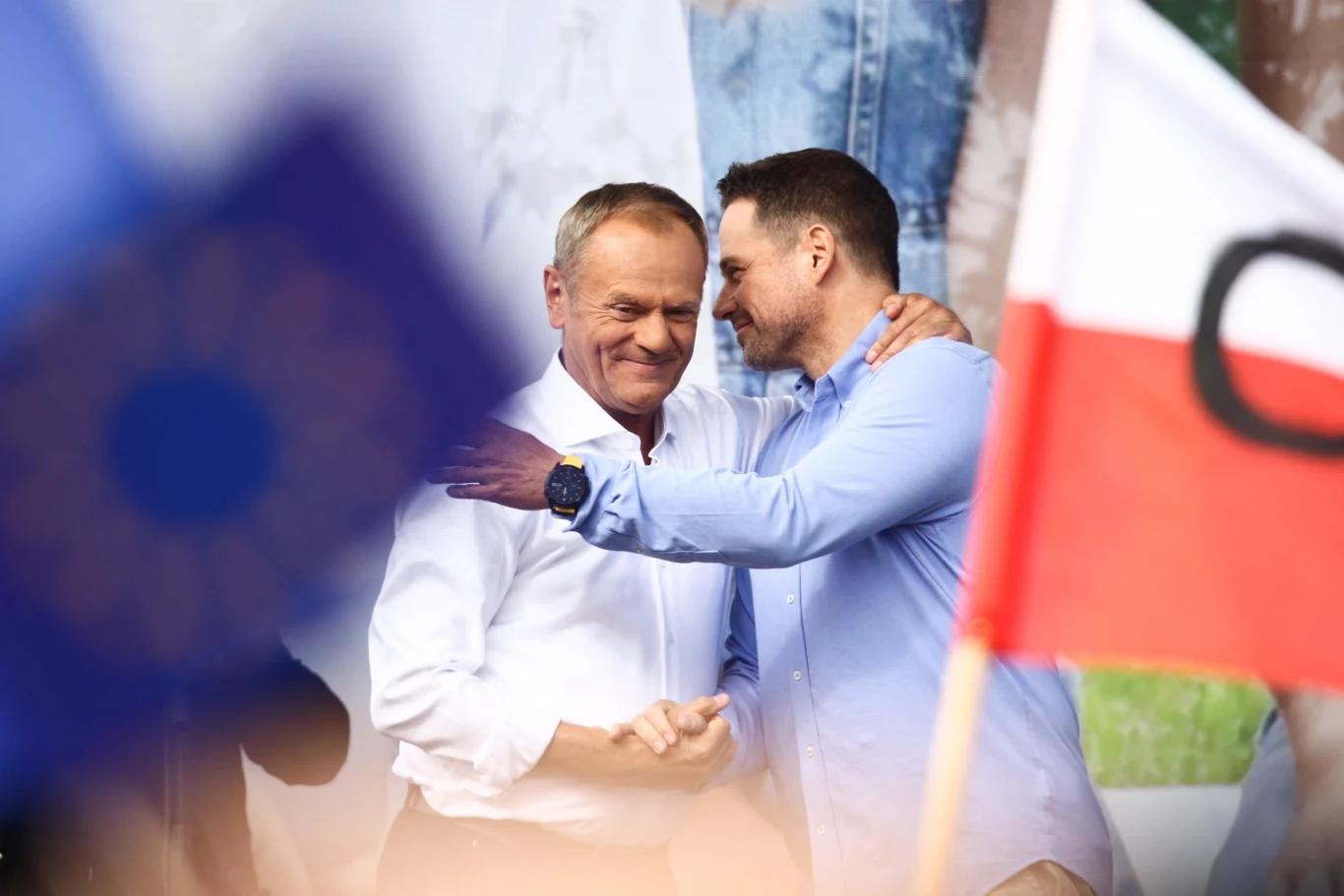 Dwóch mężczyzn obejmuje się serdecznie na tle flag Polski i Unii Europejskiej, uśmiechając się do siebie podczas wydarzenia publicznego.