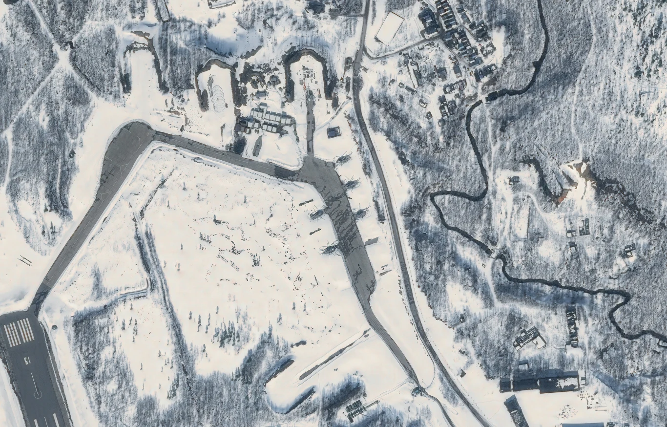 Kompleks zabudowań otoczonych lasem pokrytym śniegiem, widoczny rozbudowany układ dróg i drobnych obiektów, wyraźnie zaznaczona infrastruktura w postaci pasa startowego oraz rozproszonych konstrukcji na terenie całego obszaru.