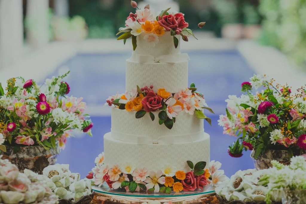 Tort weselny z prawdziwego zdarzenia, bogato zdobiony, na duże wesele, to koszt nawet 2000 zł Trzypiętrowy biały tort ozdobiony kolorowymi kwiatami z masy cukrowej, ustawiony na stole wśród bukietów świeżych kwiatów, w tle rozmazany basen lub fontanna.