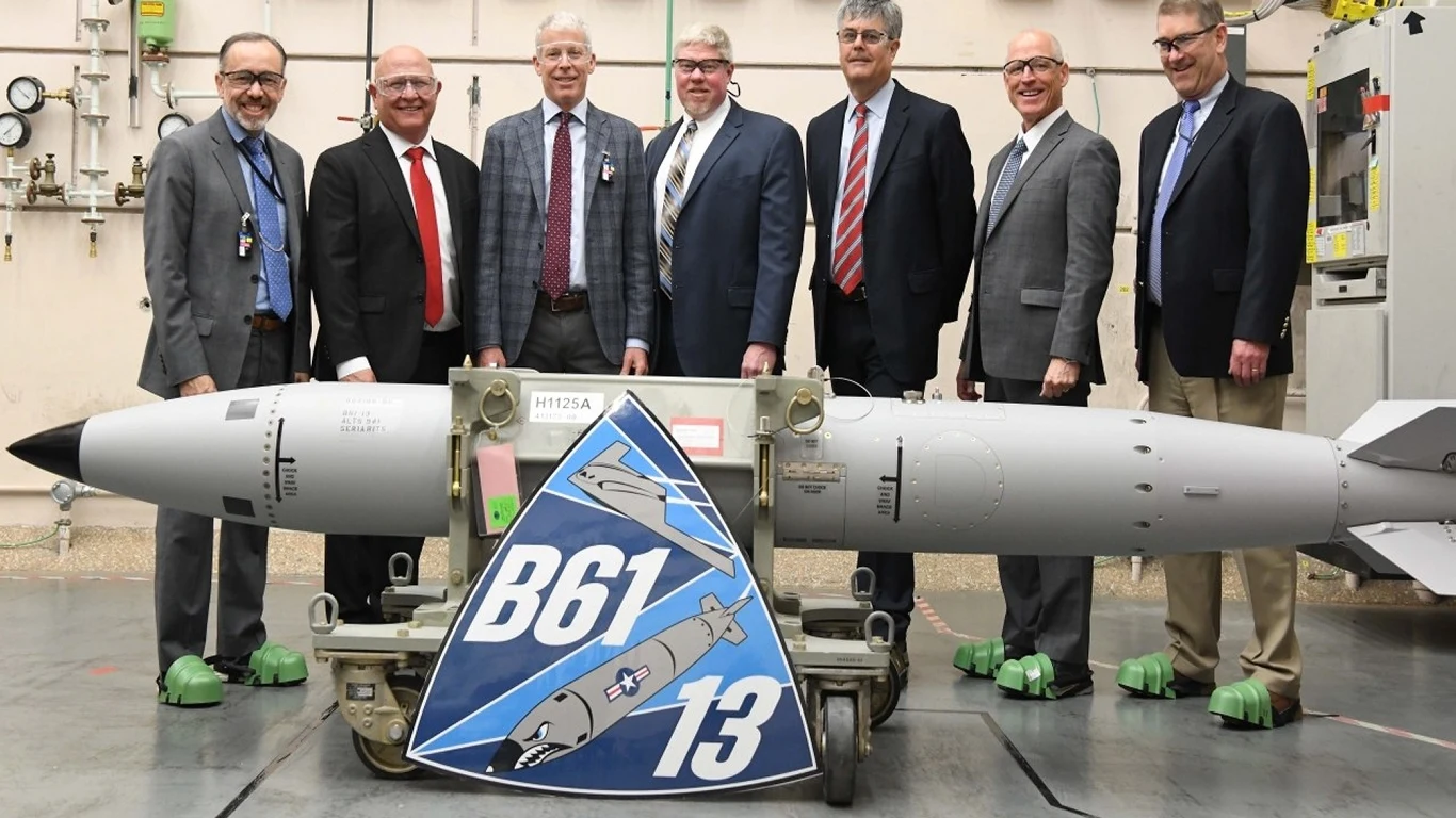 Stany Zjednoczone prezentują nową bombę atomową B61-13 Grupa siedmiu mężczyzn w garniturach pozuje wewnątrz budynku technicznego przy dużym ładunku bombowym oznaczonym jako B61-13, na stojaku znajduje się duży emblemat z napisem oraz grafiką bomby.