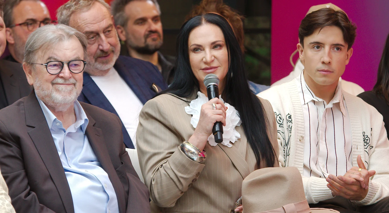 Konferencja prasowa Polsat Hit Festiwalu 2025 Trzy osoby siedzą w pierwszym rzędzie podczas wydarzenia, kobieta w środku trzyma mikrofon i mówi, otoczona przez mężczyzn w eleganckich ubraniach, w tle widoczni inni uczestnicy spotkania.