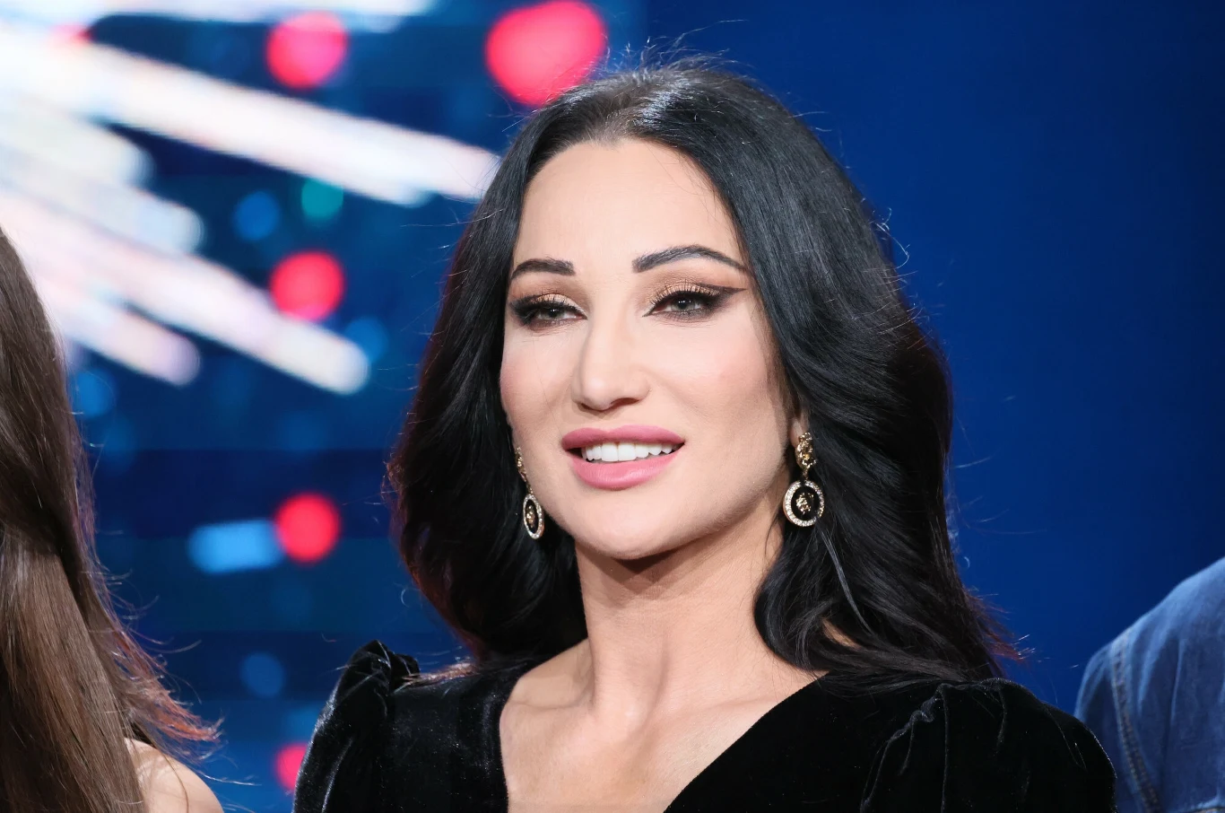 Justyna Steczkowska Uśmiechnięta kobieta o ciemnych, falowanych włosach w eleganckiej, czarnej kreacji, z efektownym makijażem i wiszącymi kolczykami, na tle rozmytych, kolorowych świateł.