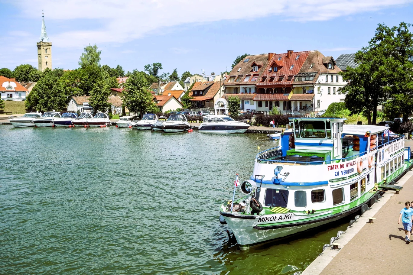 Statek wycieczkowy zacumowany przy brzegu jeziora, z tyłu widać marinę z łodziami motorowymi oraz zabytkowe, wielopiętrowe domy otoczone zielenią i wyraźnią wieżę kościelną w tle.