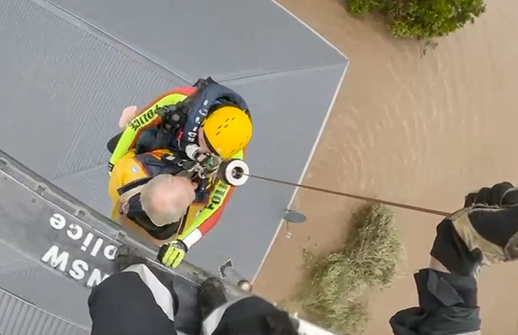 Ratownik w żółtym kasku ewakuuje osobę za pomocą wyciągarki z helikoptera, nad zalanym przez powódź obszarem; akcja odbywa się na dachu budynku z napisem 'POLICE'