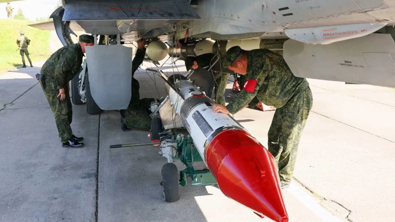 Żołnierze w mundurach przygotowują uzbrojenie pod skrzydłem wojskowego samolotu, centralnie widoczna duża bomba z czerwonym czubkiem, otoczenie to lotnisko wojskowe.