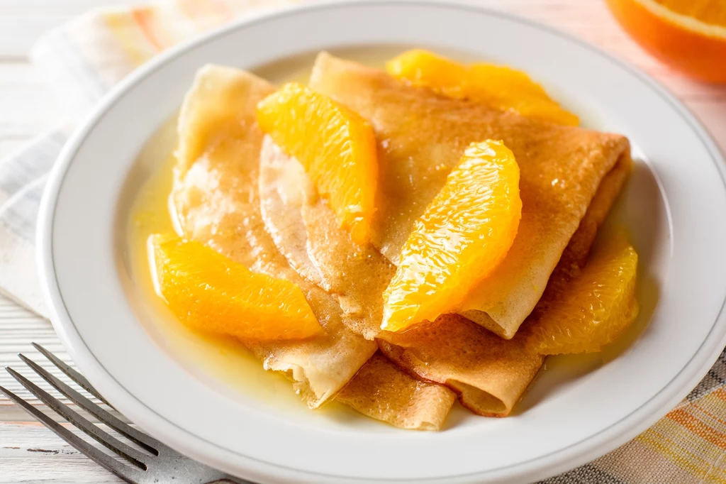 Crêpes Suzette to klasyka francuskiej kuchni i małe kulinarne widowisko Naleśniki polane sosem pomarańczowym, udekorowane kawałkami świeżych pomarańczy na białym talerzu.