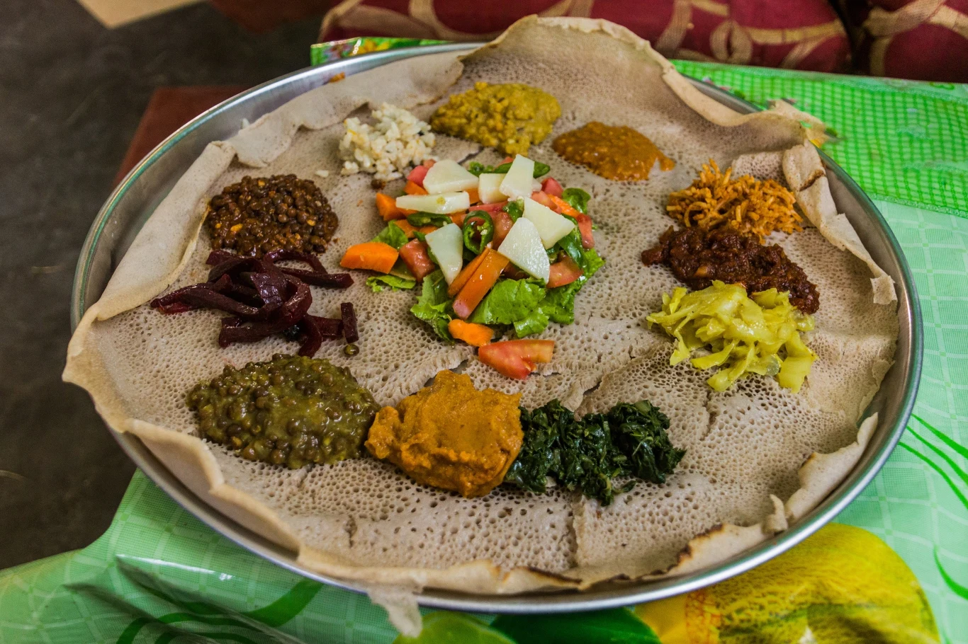 Etiopski placek injera z rozmaitymi dodatkami, w tym duszonymi warzywami, sałatką z pomidorów, marchewki i białych warzyw, a także różnymi pastami i potrawkami rozmieszczonymi wokół brzegu tacy.
