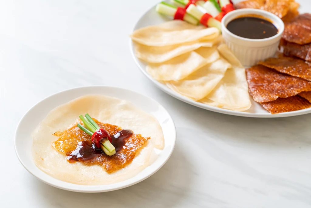 Peking duck pancakes - po zwinięciu naleśnika maczamy go w sosie Plaster chrupiącej kaczki z sosem, pokrojonym ogórkiem i cebulą na cienkim naleśniku, obok duży talerz z dodatkowymi składnikami oraz miseczka z ciemnym sosem.