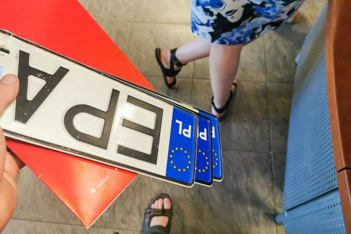 Koniec ze staniem w kolejkach w urzędzie. Starostwo wyśle tablice do Paczkomatu Trzy polskie tablice rejestracyjne z literami 'EPA' trzymane w ręce, fragmenty stóp kilku osób w sandałach oraz kawałek czerwonego blatu i podłogi.