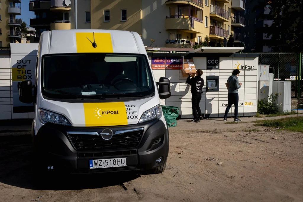 InPost wyszedł naprzeciw urzędom i ich interesantom. Samochód dostawczy InPost zaparkowany przy paczkomacie, z przodu widoczna maska pojazdu, w tle ludzie odbierający przesyłki oraz budynki mieszkalne.