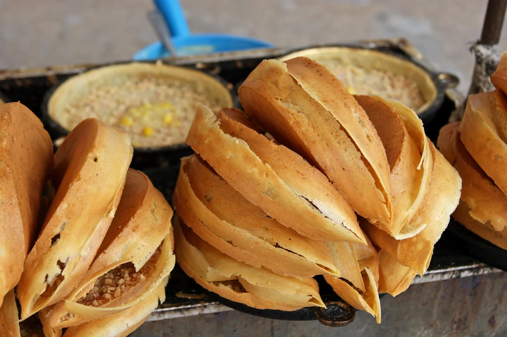 Naleśniki Apam Balik, wypełnione kukurydzą, fistaszkami i cukrem to prawdziwy malezyjski street food Stos świeżo upieczonych, chrupiących naleśników lub placków z nadzieniem orzechowym oraz foremki z ciastem w tle.