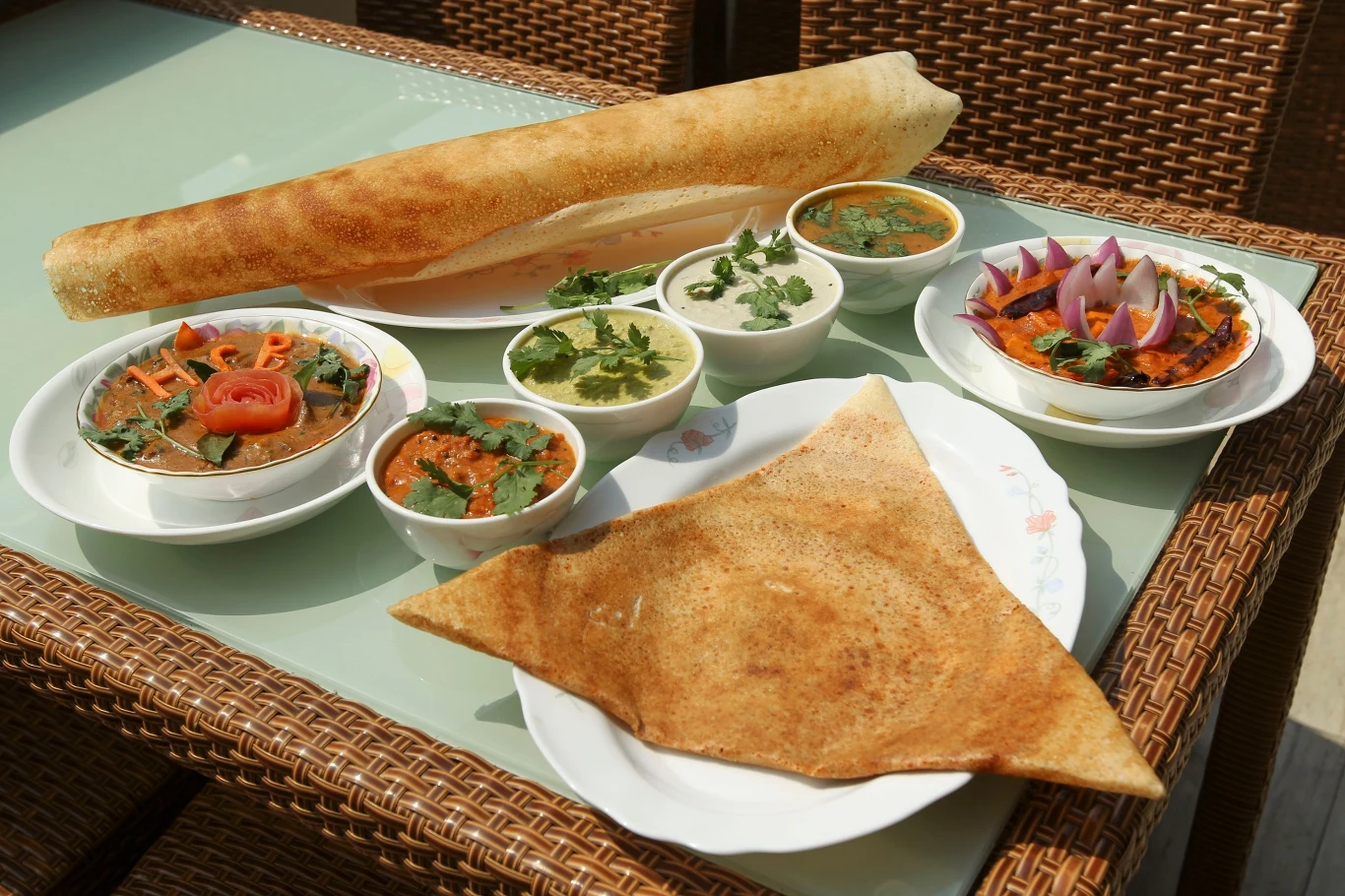 Różnorodne dania kuchni indyjskiej podane na szklanym stole, w tym chrupiące placki dosa, kilka misek z sosami, curry i chutney, udekorowane świeżymi ziołami oraz pokrojoną cebulą i pomidorem.
