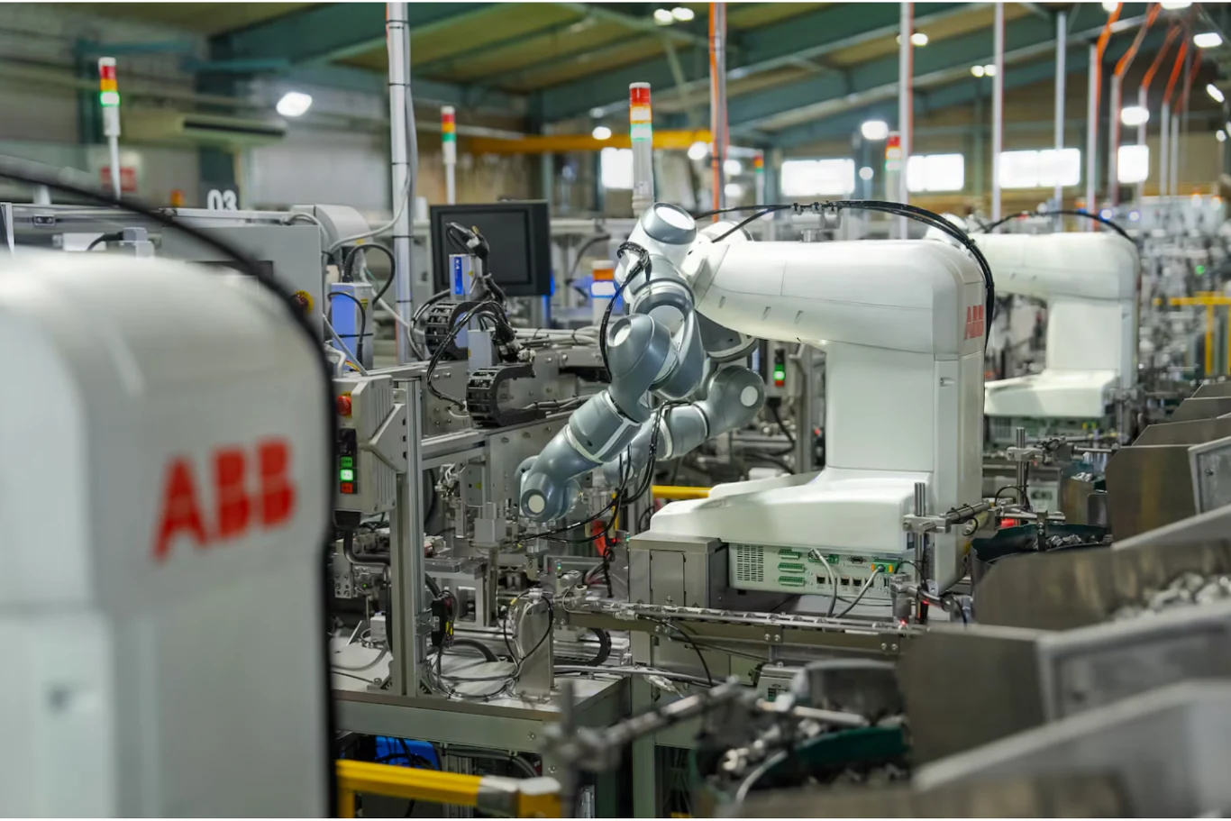 Przemysłowe roboty ABB przy taśmie produkcyjnej w nowoczesnej fabryce, z licznymi podzespołami elektronicznymi, monitorami i przewodami w uporządkowanym środowisku przemysłowym.