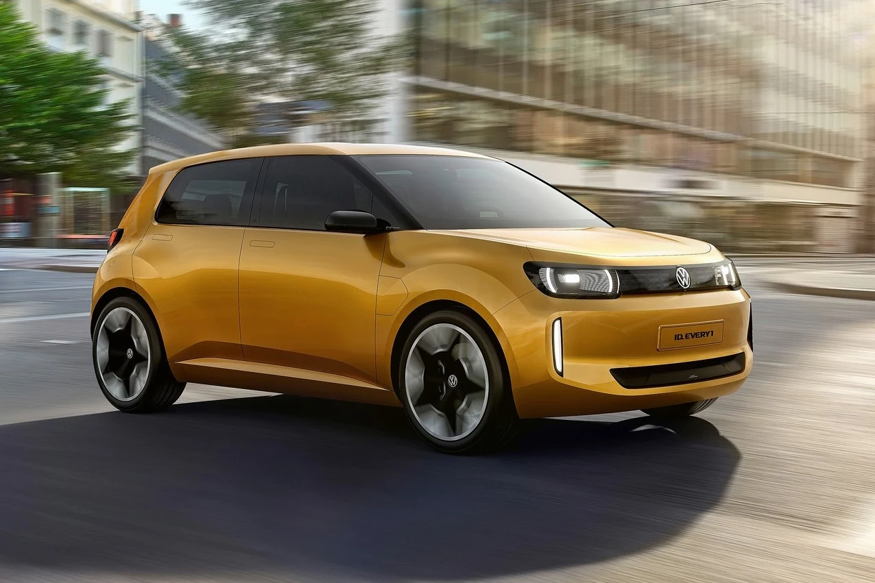 Volkswagen ID. Every1 to zapowiedź najtańszego i najmniejszego elektryka ID.1 Nowoczesny miejski samochód w kolorze złotym porusza się po ulicy w otoczeniu wysokich przeszklonych budynków i drzew.