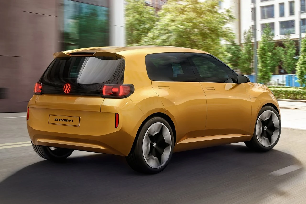 Volkswagen ID. Every1 Jasnożółty samochód osobowy marki Volkswagen poruszający się miejską ulicą, nowoczesna sylwetka auta z dużymi sportowymi felgami oraz przyciemnianymi szybami, w tle zabudowa miejska i roślinność.